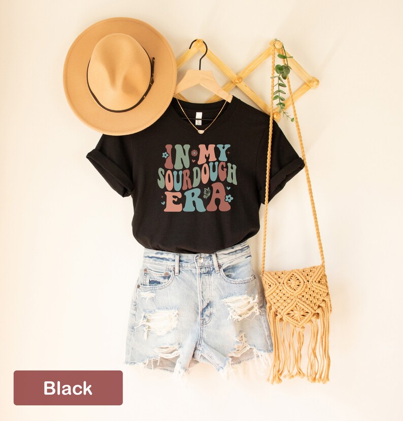 May include: Black t-shirt with the text "In My Sourdough Era" in a retro font with floral accents. The shirt is styled with denim shorts, a straw hat, and a woven bag.