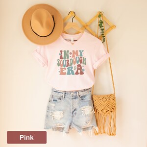 May include: Pink t-shirt with the text "In My Sourdough Era" in a retro font with floral accents. The shirt is on a hanger with a straw hat and a woven bag.