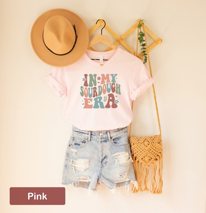 May include: Pink t-shirt with the text "In My Sourdough Era" in a retro font with floral accents. The shirt is on a hanger with a straw hat and a woven bag.