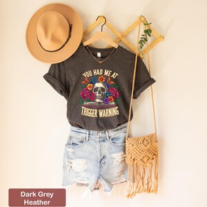 Puede incluir: Camiseta de cuello en V de color gris oscuro jaspeado con un gr&aacute;fico de calavera y flores y el texto "You had me at trigger warning".