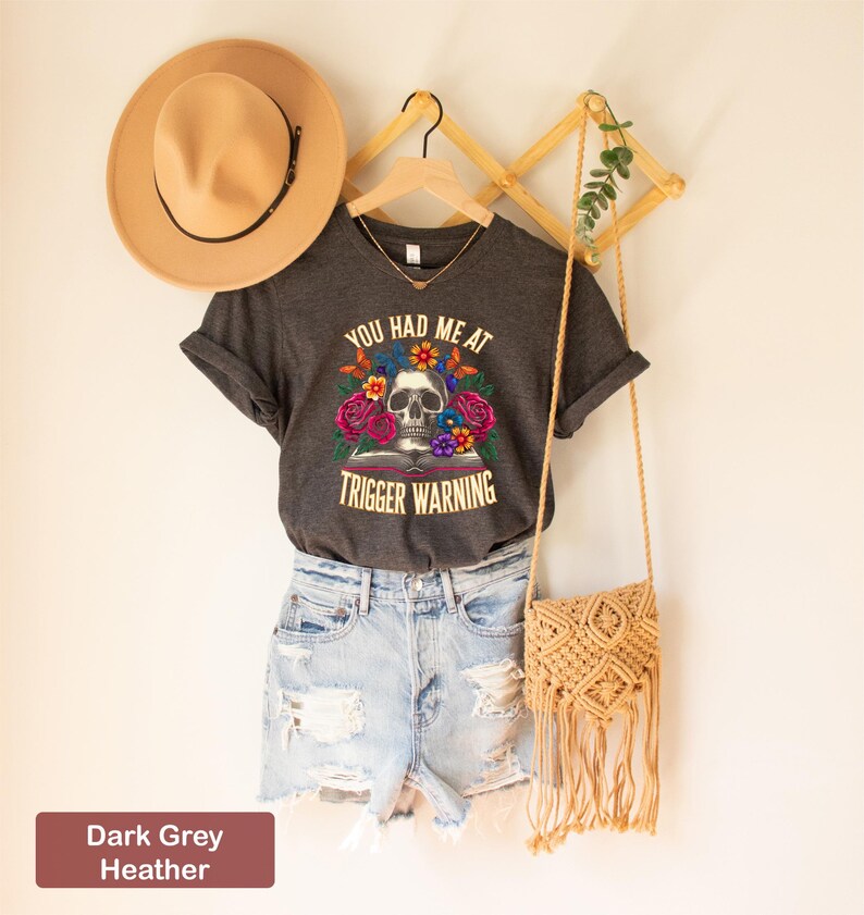 Puede incluir: Camiseta de cuello en V de color gris oscuro jaspeado con un gr&aacute;fico de calavera y flores y el texto "You had me at trigger warning".