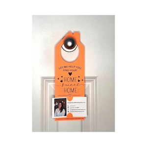 May include: Orange door hanger with a house-shaped top and a circular opening. The text "LET ME HELP YOU FIND YOUR HOME sweet HOME" is printed on the front. A business card with a photo and contact information is attached.