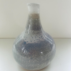 May include: A handcrafted ceramic vase with a bulbous body and a narrow neck. The vase features a speckled, textured surface with a gradient of colors, transitioning from white at the top to shades of blue and gray at the base. A decorative home decor item.