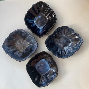 May include: Four handmade ceramic bowls with a unique, organic shape. The bowls feature a dark blue glaze with lighter blue and gray accents, creating a marbled effect. The bowls are arranged in a diamond pattern, showcasing their individual designs.