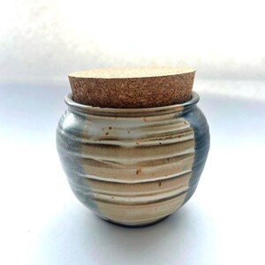 May include: A small, round ceramic jar with a ribbed texture and a natural cork lid. The jar features a glaze with horizontal bands of cream, tan, and dark blue, with subtle brown speckles visible across the surface of the clay body.