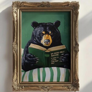 Op de afbeelding: Een ingelijst kunstwerk toont een zwarte beer die een groen boek leest met de titel "Un-Bear-Ably Awesome: A Guide to Loving Your Fuzzy Self". De beer zit op een gestreept kussen tegen een groene achtergrond. De lijst is goudkleurig en versierd.