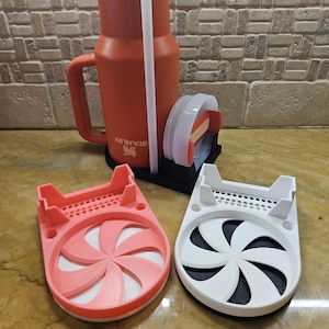 Puede incluir: Dos posavasos impresos en 3D con un diseño de espiral. Un posavasos es rosa y el otro es blanco. Los posavasos están sobre una encimera.