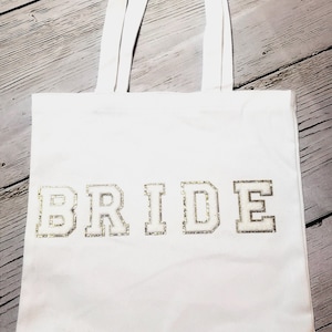 May include: White tote bag with the word "BRIDE" in gold glitter lettering. The bag has two white handles and is made of a sturdy fabric. Perfect for carrying essentials.