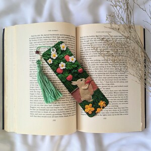 May include: A handmade bookmark featuring a green and brown clay rabbit eating a strawberry. The bookmark is decorated with a green and white clay strawberry plant with red strawberries and white flowers. The bookmark has a green tassel.