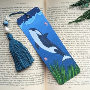 May include: A teal blue bookmark with a tassel features a hand-painted illustration of an orca swimming through a blue ocean with green seaweed and jellyfish. The bookmark is made of paper and has a slightly textured surface.