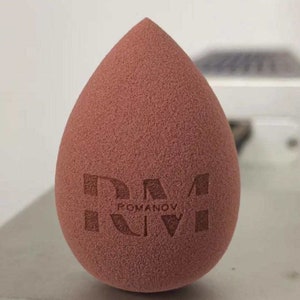 May include: A brown teardrop-shaped makeup sponge with the text "ROMANOV" printed on it.