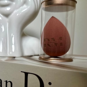 May include: A brown makeup sponge in a clear plastic container with a gold lid. The sponge has the text "RM ROUVANO" printed on it.