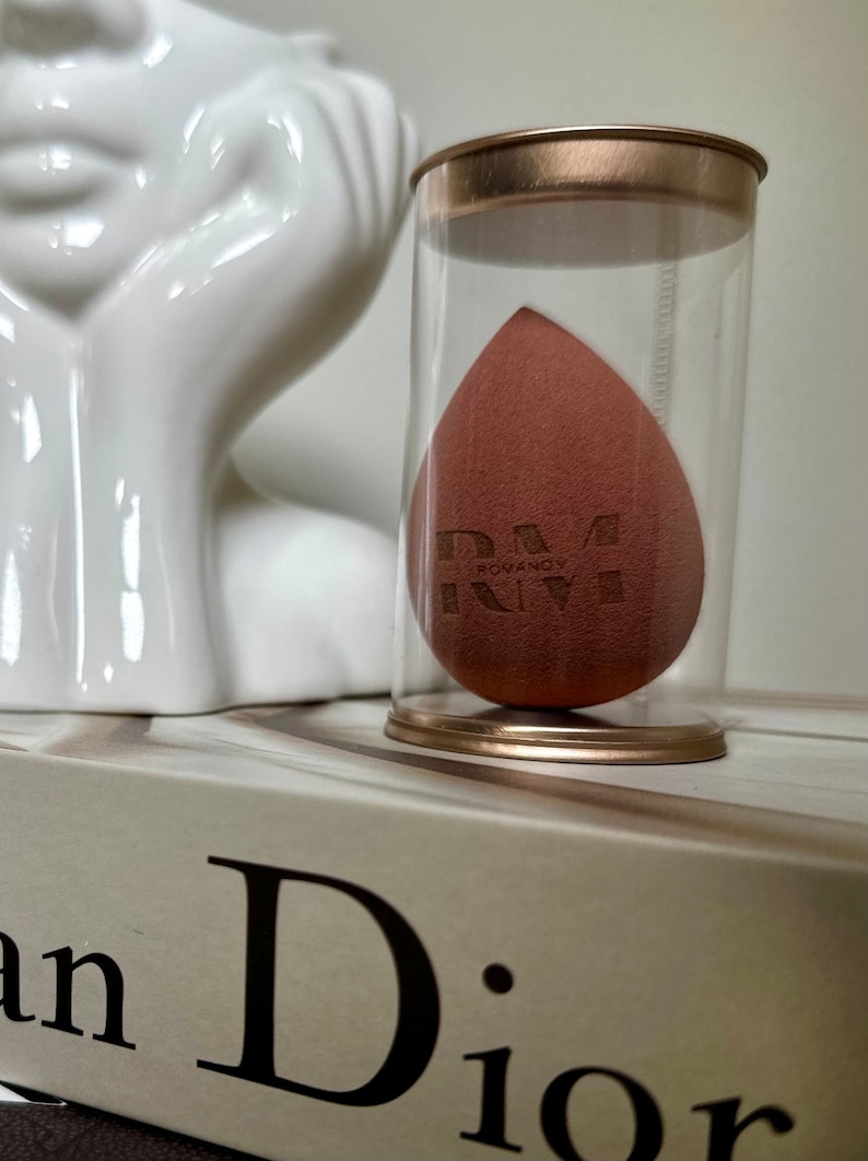 May include: A brown makeup sponge in a clear plastic container with a gold lid. The sponge has the text "RM ROUVANO" printed on it.