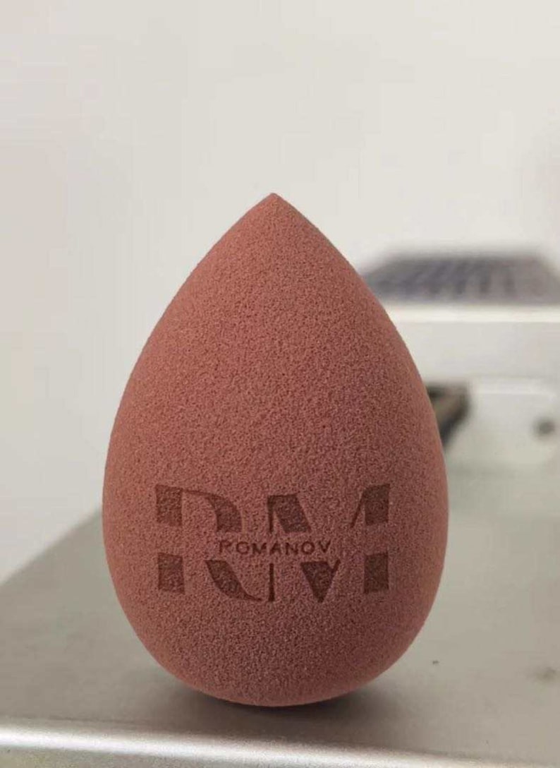 May include: A brown teardrop-shaped makeup sponge with the text "ROMANOV" printed on it.