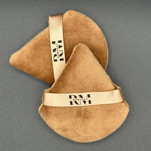 May include: Two tan, triangular powder puffs with a soft, velvety texture. Each puff has a beige ribbon with the black text "RM ROMANOV". The puffs are arranged on a gray background.