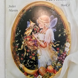 May include: A book cover titled "Bumble Babies Bee Attitudes Book 1" by Juliet Martin. The cover features a painting of a fairy with a bee, holding a basket of fruit. The artwork is set within an oval frame with a gold border, against a dark background.