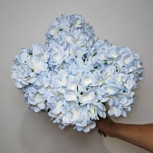 Pu&ograve; includere: Un mazzo di fiori di ortensia artificiali di colore azzurro chiaro. I fiori sono disposti a grappolo e hanno un aspetto delicato e realistico.