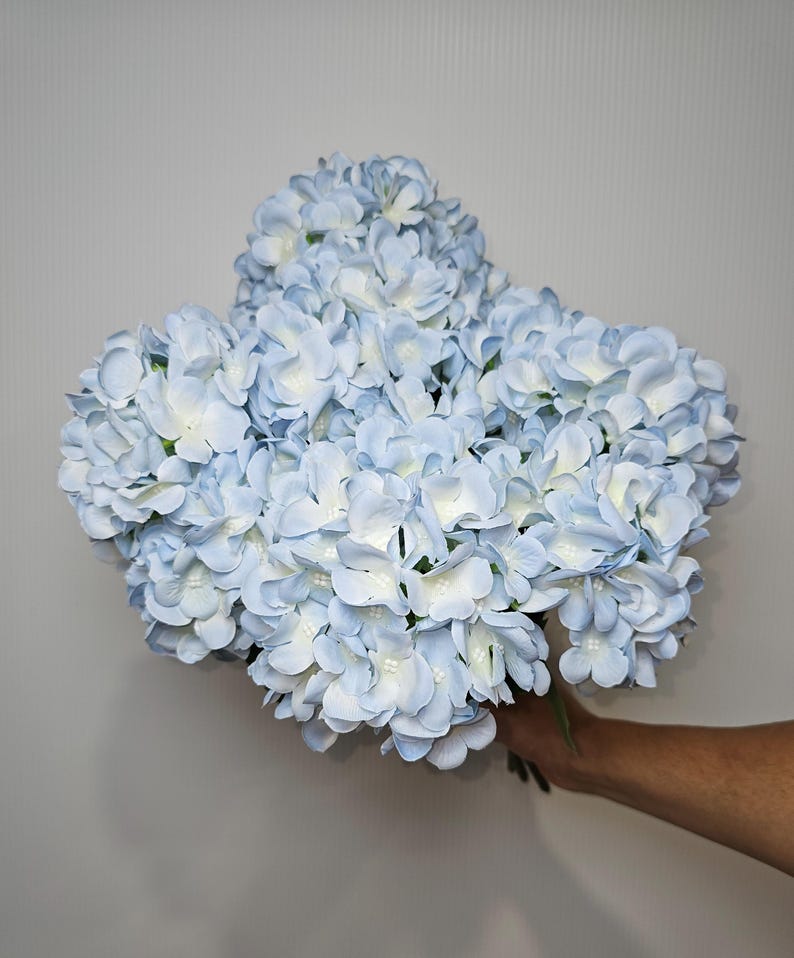 Pu&ograve; includere: Un mazzo di fiori di ortensia artificiali di colore azzurro chiaro. I fiori sono disposti a grappolo e hanno un aspetto delicato e realistico.