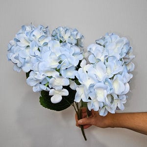 Pu&ograve; includere: Un mazzo di fiori di ortensia artificiali di colore azzurro chiaro con foglie verdi.
