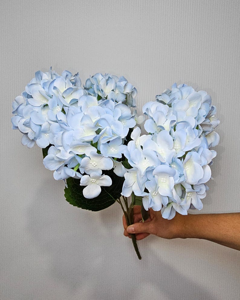 Pu&ograve; includere: Un mazzo di fiori di ortensia artificiali di colore azzurro chiaro con foglie verdi.