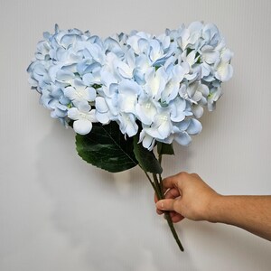Pu&ograve; includere: Un mazzo di fiori di ortensia artificiali di colore azzurro chiaro con foglie verdi.