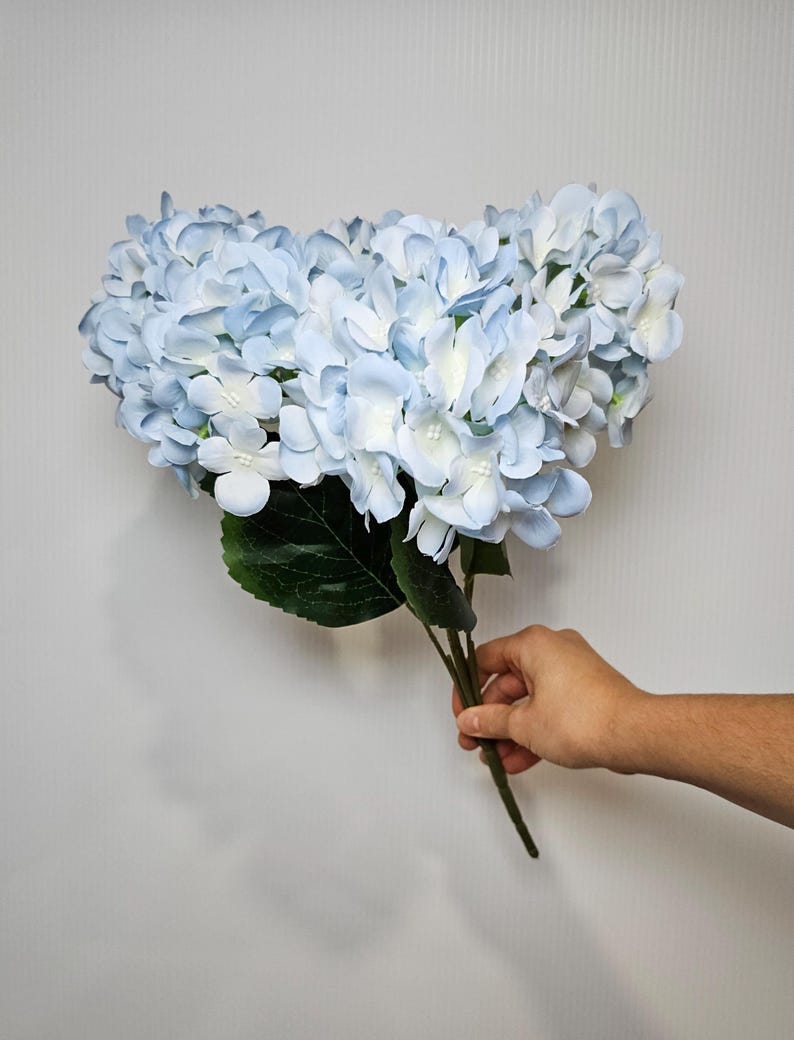 Pu&ograve; includere: Un mazzo di fiori di ortensia artificiali di colore azzurro chiaro con foglie verdi.