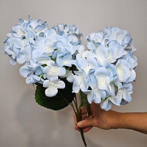 Pu&ograve; includere: Un mazzo di fiori di ortensia artificiali di colore azzurro chiaro con foglie verdi.
