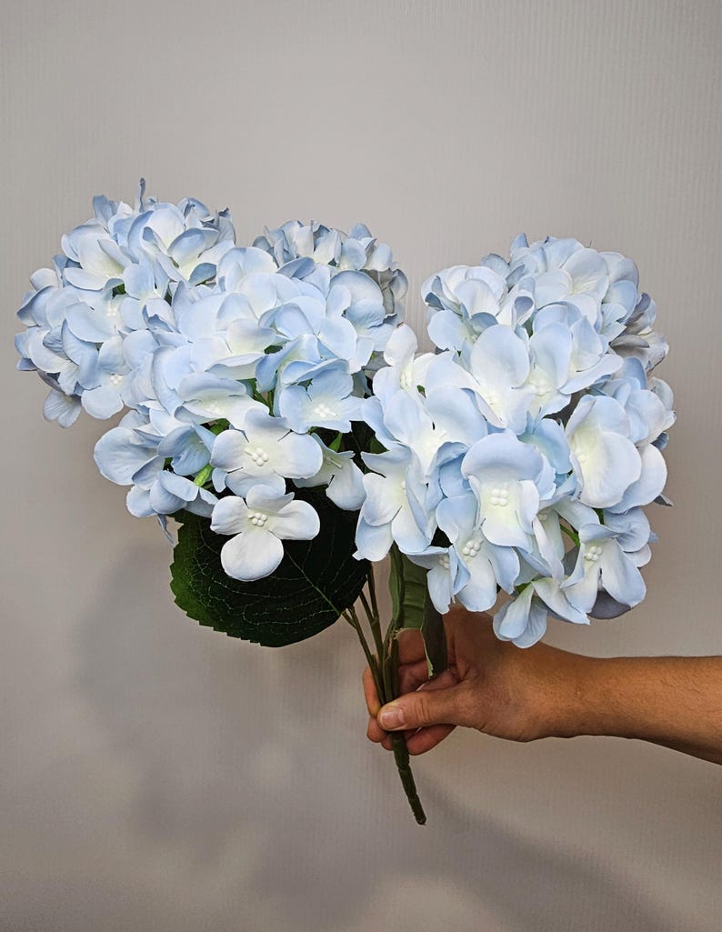 Pu&ograve; includere: Un mazzo di fiori di ortensia artificiali di colore azzurro chiaro con foglie verdi.