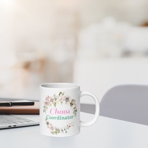 Könnte beinhalten: Weiße Keramiktasse mit Blumendesign und dem Text "Chaos Coordinator" in rosa und grüner Schrift.