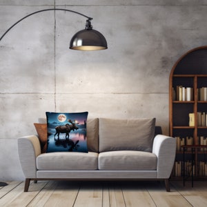 May include: A gray couch with two pillows, one with a brown and white pattern and the other with a moose standing in a lake under a full moon. The couch is in front of a gray wall with a black floor lamp.