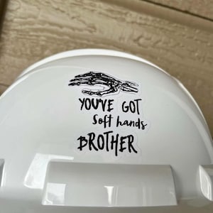 May include: White hard hat with a black and white sticker that says "You've got soft hands brother". The sticker features a skeleton hand.