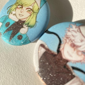 May include: Two oval pendants with anime-style illustrations on a glittery blue background. One shows a character with green hair and a blue outfit, the other a character with white hair and a brown outfit. The pendants have a sparkly finish.