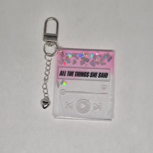 May include: A clear keychain with a pink top section and the text "ALL THE THINGS SHE SAID". The keychain has a silver clasp and chain with a heart charm. The design resembles a music player interface.