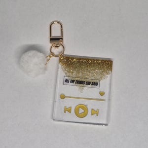 May include: Clear acrylic keychain with gold glitter top and gold hardware. The keychain features a music player design with the text "ALL THE THINGS SHE SAID" and a white pom-pom charm.