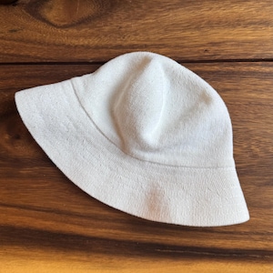 May include: A solid off-white bucket hat featuring a knit construction with a rounded crown and a downward-sloping brim. The hat is crafted from a textured fabric with a consistent weave pattern throughout the entire surface of the headwear.