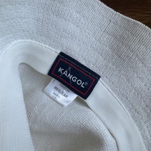 May include: A white textured bucket hat featuring a dark blue rectangular label with the white text KANGOL and a red border. A white fabric tag underneath displays the text 1781CL01, REGULAR, and MADE IN UK, indicating the product details clearly.