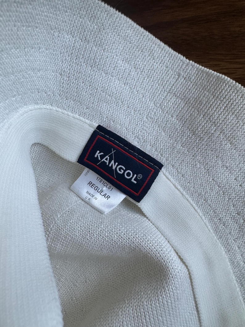 May include: A white textured bucket hat featuring a dark blue rectangular label with the white text KANGOL and a red border. A white fabric tag underneath displays the text 1781CL01, REGULAR, and MADE IN UK, indicating the product details clearly.