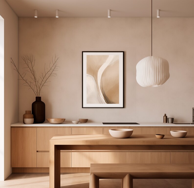 May include: A modern kitchen with light wood cabinetry and a matching island. A framed abstract art piece hangs on the wall. A white, textured pendant light hangs above the island. Decorative bowls and a vase with branches add to the decor.
