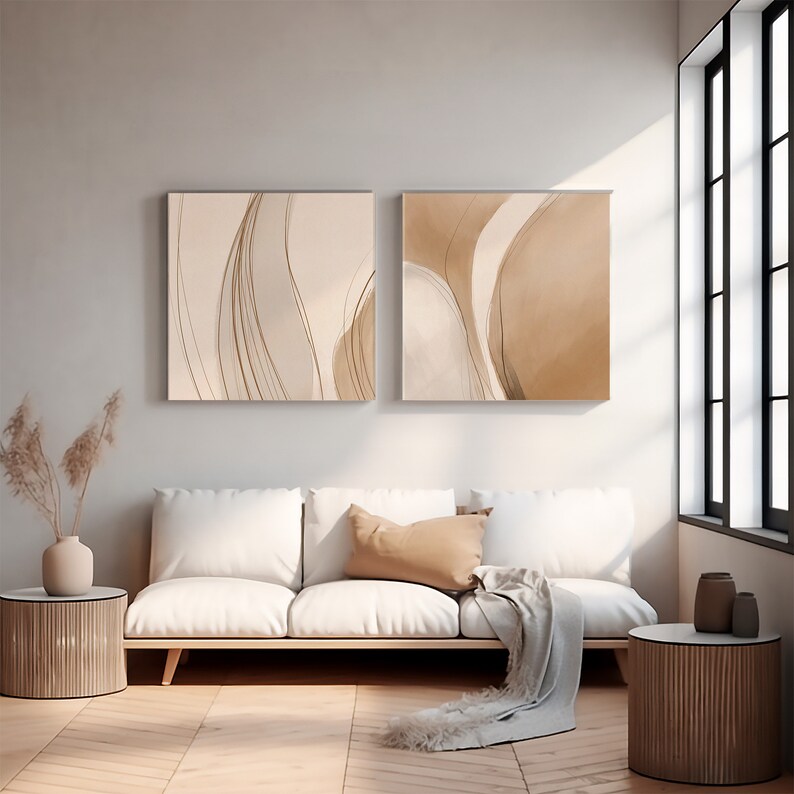May include: Two abstract art prints with beige and brown tones hang above a white sofa. The prints feature flowing lines and shapes. A tan throw pillow and a light gray blanket rest on the sofa. The room has a modern, minimalist aesthetic.