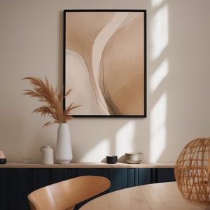 May include: Abstract art print in a black frame. The artwork features a watercolor design with beige and brown tones and thin black lines. A white vase with dried pampas grass sits below the artwork, with a wooden chair and table in the foreground.