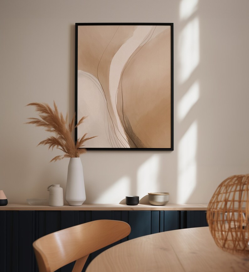 May include: Abstract art print in a black frame. The artwork features a watercolor design with beige and brown tones and thin black lines. A white vase with dried pampas grass sits below the artwork, with a wooden chair and table in the foreground.
