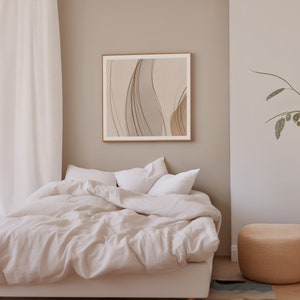 May include: A framed abstract art print with a beige and brown color palette hangs above a bed with white bedding. The room has a neutral color scheme with a beige wall and a woven ottoman.