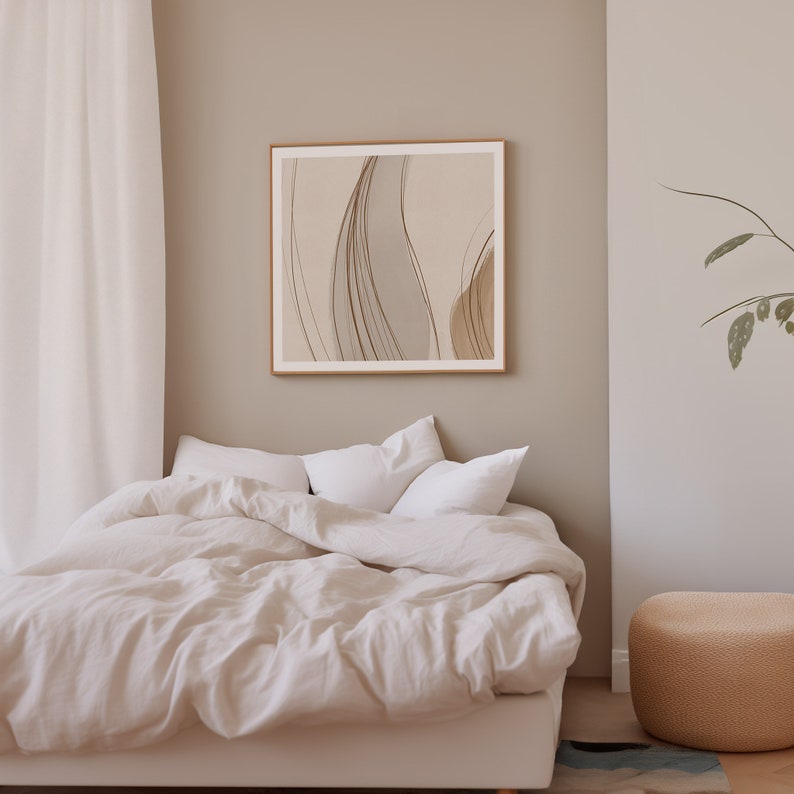 May include: A framed abstract art print with a beige and brown color palette hangs above a bed with white bedding. The room has a neutral color scheme with a beige wall and a woven ottoman.
