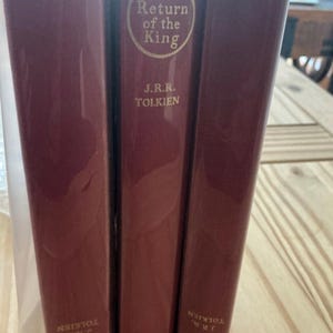 Puede incluir: Tres libros de tapa dura color granate de J.R.R. Tolkien, incluyendo The Return of the King, The Two Towers y The Fellowship of the Ring. Cada lomo presenta letras doradas y el nombre de la editorial George Allen and Unwin en vertical.