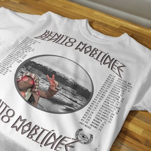 May include: White t-shirt with a black and white photo of a person wearing a bandana and sunglasses. The photo is in a circle with the text "BENITO MARTINEZ" above and below the circle. The text is in a gray font. The t-shirt also has a list of tour dates printed on the back.