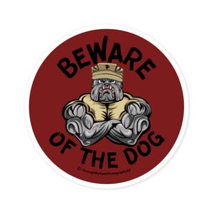 May include: A red circular sticker with a white border featuring a cartoon bulldog wearing a military hat. The bulldog has a muscular build and is standing with its arms crossed. The text "BEWARE OF THE DOG" is written in black around the bulldog.