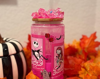 May include: A pink jar with a gold rim, filled with pink liquid and topped with pink crystals. The jar features Jack Skellington and Sally from The Nightmare Before Christmas. The background includes fall leaves and a black and white striped container.