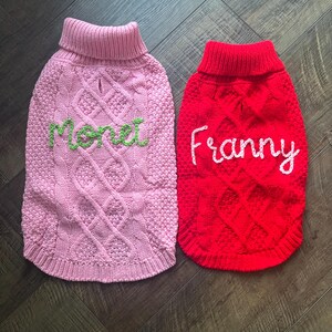 K&ouml;nnte beinhalten: Zwei gestrickte Hunde-Pullover, einer rosa mit dem Namen "Monet" in gr&uuml;n gestickt, und einer rot mit dem Namen "Franny" in wei&szlig; gestickt.