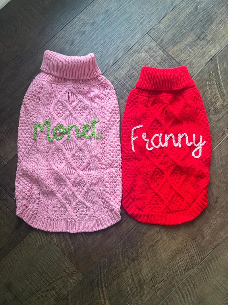 K&ouml;nnte beinhalten: Zwei gestrickte Hunde-Pullover, einer rosa mit dem Namen "Monet" in gr&uuml;n gestickt, und einer rot mit dem Namen "Franny" in wei&szlig; gestickt.