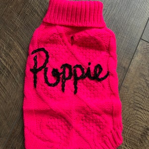 K&ouml;nnte beinhalten: Ein rosa gestrickter Hundepullover mit einem schwarzen Zopfmuster und dem Namen "Poppy" auf der Vorderseite gestickt.
